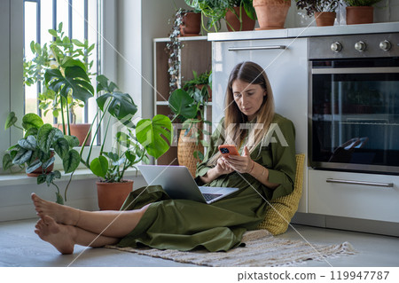 Interested woman with laptop on knees holding smartphone sitting on floor among houseplants at home. Interested woman with laptop on knees holding smartphone sitting on floor among houseplants at home. 119947787