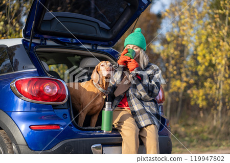 Woman pet owner drinking tea sitting in car trunk with dog enjoying nature in forest on weekend. 119947802