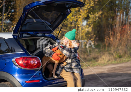 Gentle woman hugging beloved dog tightly with softness fondness while sitting in trunk of blue car. 119947882