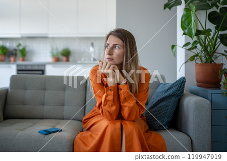Worried stressed woman feel tension sitting on sofa looking aside waiting call on cellphone at home. 119947919