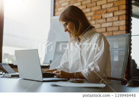 A Professional Woman Diligently Working on Her Laptop in a Contemporary Office Setting A Professional Woman Diligently Working on Her Laptop in a Contemporary Office Setting 119948448