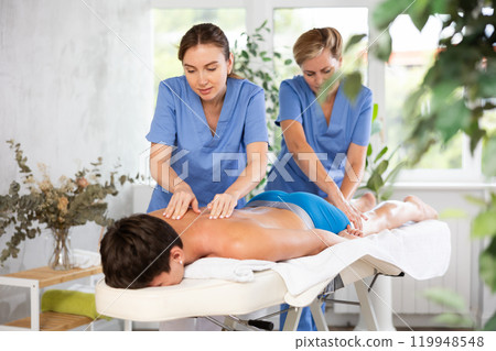 Two female masseur doing massage at back man who lying on massage table in beauty salon Two female masseur doing massage at back man who lying on massage table in beauty salon 119948548