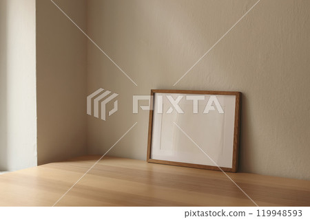 Horizontal wooden thin picture frame, empty poster mockup on wooden table near window. Beige wall background. Modern moody interior decor. Side view, copy space, no people. Minimal composition. 119948593