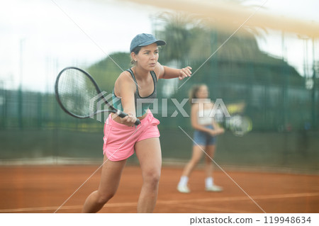 Fit girl engrossed in doubles tennis game on clay court Fit girl engrossed in doubles tennis game on clay court 119948634