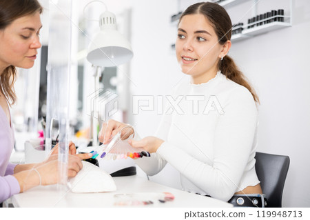 Young female manicurist showing palette of nail varnishes to woman client 119948713