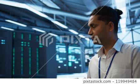 Software developer walking in data center, managing and maintaining databases, ensuring data integrity and security. Close up of man updating server room mainframe systems using tablet, camera B Software developer walking in data center, managing and maintaining databases, ensuring data integrity and security. Close up of man updating server room mainframe systems using tablet, camera B 119949484