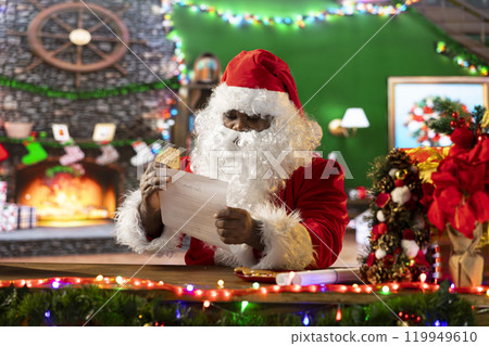 Father Christmas reading letters by the fire in his rustic log cabin, preparing presents from the North Pole workshop. Santa surrounded by festive decorations and x mas lights for seasonal cheer. 119949610