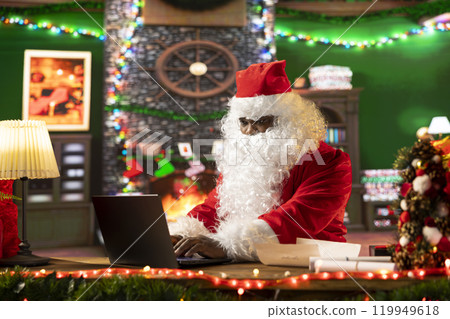 Father Christmas is navigating the web checking on children wish lists using his laptop. Festive North Pole cabin is filled with holiday cheer and online activity this x mas, checking the world map. 119949618
