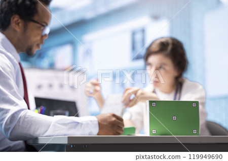 Patient and medical specialist at a check up visit next to green screen on medicine box, discussing about treatment and medical drugs for recovery. Female doctor pointing at medicaments. 119949690