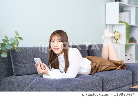 A young woman in casual clothes lying on a sofa, living room, smartphone 119950484