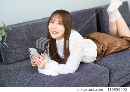 A young woman in casual clothes lying on a sofa, living room, smartphone 119950490