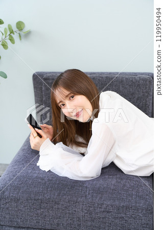 A young woman in casual clothes lying on a sofa, living room, smartphone A young woman in casual clothes lying on a sofa, living room, smartphone 119950494