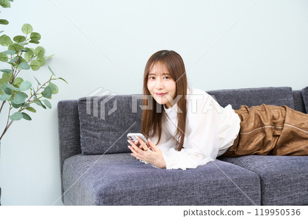 A young woman in casual clothes lying on a sofa, living room, smartphone 119950536