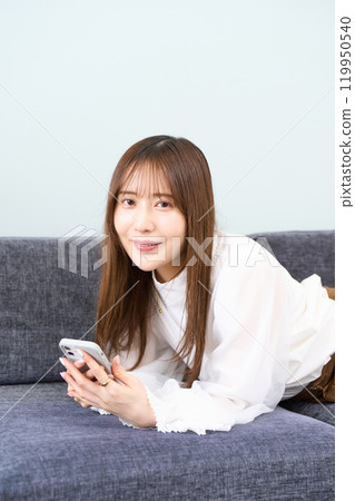A young woman in casual clothes lying on a sofa, living room, smartphone 119950540