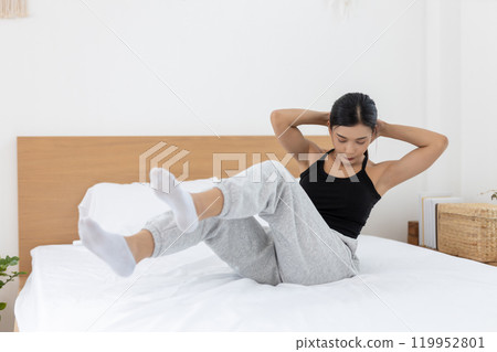Asian young woman morning exercises after waking up on bed, Stretch feet forward. Relax and stretch and workout at home in holiday. Activity body movement arm leg. 119952801