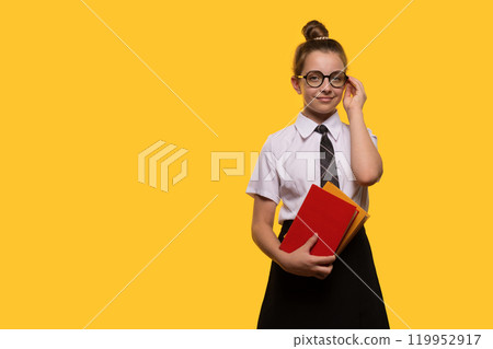 Teen girl adjusting glasses, holding books, wearing school uniform 119952917