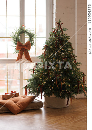 A decorated Christmas tree stands in front of a window in a cozy, bright living room A decorated Christmas tree stands in front of a window in a cozy, bright living room 119954276