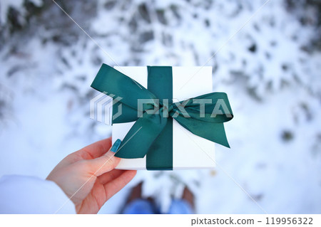 a pair of hands holding a beautifully wrapped gift box adorned with a vibrant green ribbon, set against a picturesque snowy backdrop. 119956322