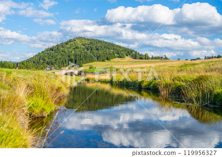 A serene view of the Jizerka village showcases the reflection of Bukovec Mountain in the calm waters of a river. Lush greenery and gentle hills surround the picturesque landscape under a cloudy sky. 119956327