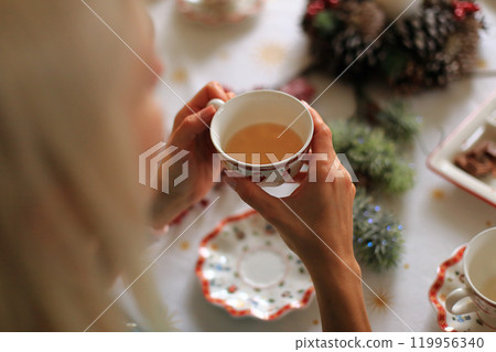a pair of hands gently holding a steaming cup of tea, set against the backdrop of a beautifully decorated Christmas table 119956340