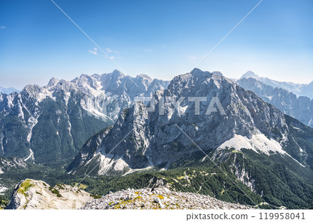 The majestic Skrlatica, Prisank, and Triglav mountains rise dramatically in the Julian Alps, showcasing rugged terrain and clear blue skies, perfect for hiking and exploring nature's beauty. The majestic Skrlatica, Prisank, and Triglav mountains rise dramatically in the Julian Alps, showcasing rugged terrain and clear blue skies, perfect for hiking and exploring nature's beauty. 119958041