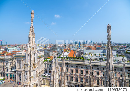 From the rooftop of the Duomo, breathtaking views of Milan's skyline reveal historic architecture and modern buildings under a clear sky. The intricate spires stand tall, showcasing the city's charm. 119958073
