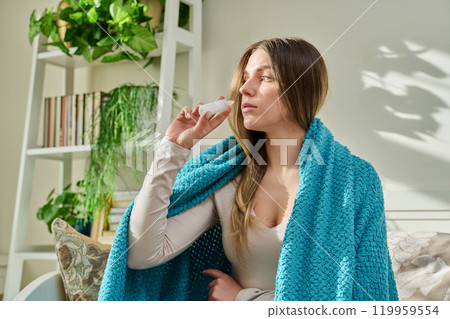 Young woman with runny nose using nasal spray sitting on couch at home. 119959554