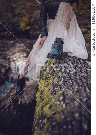 A woman in a flowing skirt sits on a moss-covered log by a tranquil stream in the forest 119961197
