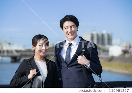 兩個新鮮的男女,看起來像新推銷員,穿著西裝站在河邊,看著鏡頭 兩個新鮮的男女,看起來像新推銷員,穿著西裝站在河邊,看著鏡頭 119961237