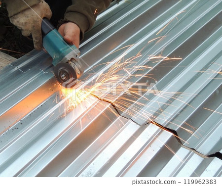 workers' hands in one glove hold a grinder with a sharp rotating disk and cut a sheet of silvery thin relief iron with splashes of bright sparks workers' hands in one glove hold a grinder with a sharp rotating disk and cut a sheet of silvery thin relief iron with splashes of bright sparks 119962383