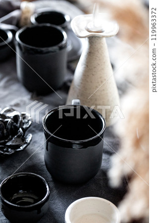 Rustic table setting with empty craft handmade ceramic tableware, black and white rough bowls, cups, jug and vase on linen tablecloth. Dry reed flowers. Day light dining room. 119963372