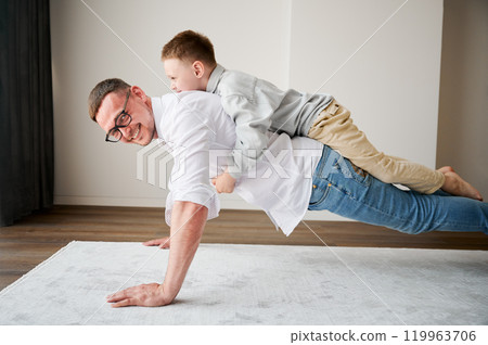 Energetic father and his little son training at home. Kid climbed dad's back while he doing plank. Young family enjoying activity games at home. Energetic father and his little son training at home. Kid climbed dad's back while he doing plank. Young family enjoying activity games at home. 119963706