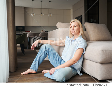Portrait of woman sitting next to couch in her apartment. Pretty female in casual clothes looking away. 119963710