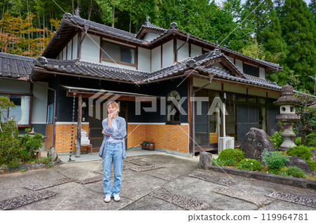 A woman tilts her head in front of an old house, vacant house, inheritance, countryside 119964781