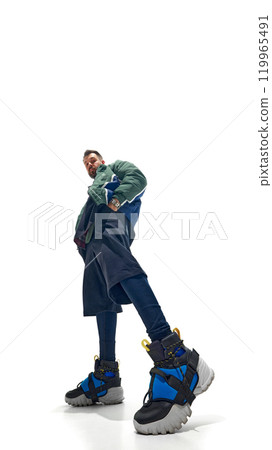 Low-angle shot of young man in layered outfit, with colorful sneakers and green bomber jacket against white studio background. 119965491