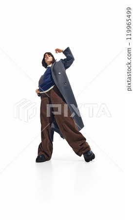 Young woman in oversized grey coat and wide brown pants strikes dynamic pose against white studio background, embodying blend of youthful energy and urban fashion Young woman in oversized grey coat and wide brown pants strikes dynamic pose against white studio background, embodying blend of youthful energy and urban fashion 119965499