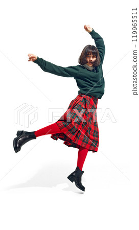 Energetic woman in green sweater and plaid skirt jumps with arms raised, embodying joy and freedom in urban fashion against white studio background. Energetic woman in green sweater and plaid skirt jumps with arms raised, embodying joy and freedom in urban fashion against white studio background. 119965511