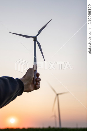 Man holds toy wind turbine in his hand. Real wind turbines in the background 119966599