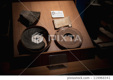 Antique film reels and Kinora projector discs displayed in a dimly lit storage box at a historical exhibit in London. Old film reels and Kinora projector discs are arranged neatly in a dark wooden box 119968341