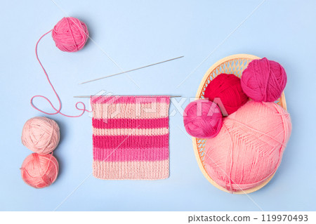 Pink and magenta knitting balls in basket with knitting needles and knitted scarf on blue background. Set for the hobby of knitting warm clothes or toys. Concept needlework Pink and magenta knitting balls in basket with knitting needles and knitted scarf on blue background. Set for the hobby of knitting warm clothes or toys. Concept needlework 119970493