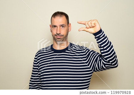 Bearded hispanic man in his 40s wearing a striped sweater with an annoyed expression making a size sign with his hand with his fingers while looking at the camera. Measurement concept Bearded hispanic man in his 40s wearing a striped sweater with an annoyed expression making a size sign with his hand with his fingers while looking at the camera. Measurement concept 119970928