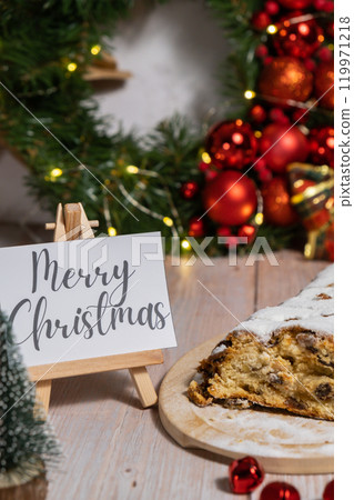 Note with saying MERRY CHRISTMAS congratulations. Traditional German Christmas stollen cake in background of handmade festive decor. Winter holiday treats. Vegan baking recipe 119971218