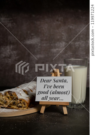 Paper note with text DEAR SANTA I HAVE BEEN GOOD ALMOST ALL YEAR joke humor pun.Homemade Christmas traditional cake pie with marzipan nuts and dried fruits pastry dessert stollen with glass of milk 119971224