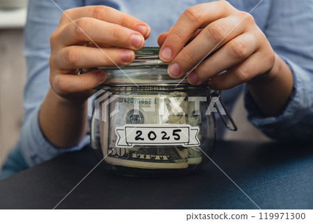 Female hands holding Glass jar full of American currency dollars cash banknote with text 2025 year. Preparation saving money. Moderate consumption and economy Female hands holding Glass jar full of American currency dollars cash banknote with text 2025 year. Preparation saving money. Moderate consumption and economy 119971300