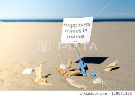 HAPPINESS COMES IN WAVES text on paper greeting card in anchor paper holder and starfish seashell summer vacation decor. Sandy beach sun coast. Holiday concept postcard. Getting away Travel 119971336