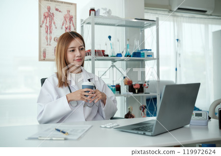 A woman in a white lab coat is sitting at a desk with a laptop 119972624