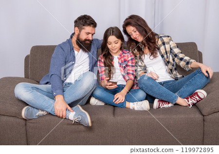 Happy father and mother look at girl child chatting in smartphone sitting on sofa, chat Happy father and mother look at girl child chatting in smartphone sitting on sofa, chat 119972895