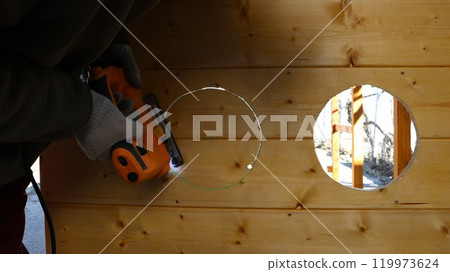 A builder wearing gloves cuts a circle according to the markings from a wooden wall using an electric jigsaw. 119973624