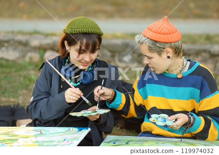 two young and innovative female artists conducting an outdoor painting lesson. Surrounded by nature, they are armed with easels, brushes, and a palette of colors, inspiring each other and their 119974032
