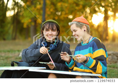 two young and innovative female artists conducting an outdoor painting lesson. Surrounded by nature, they are armed with easels, brushes, and a palette of colors, inspiring each other and their 119974033